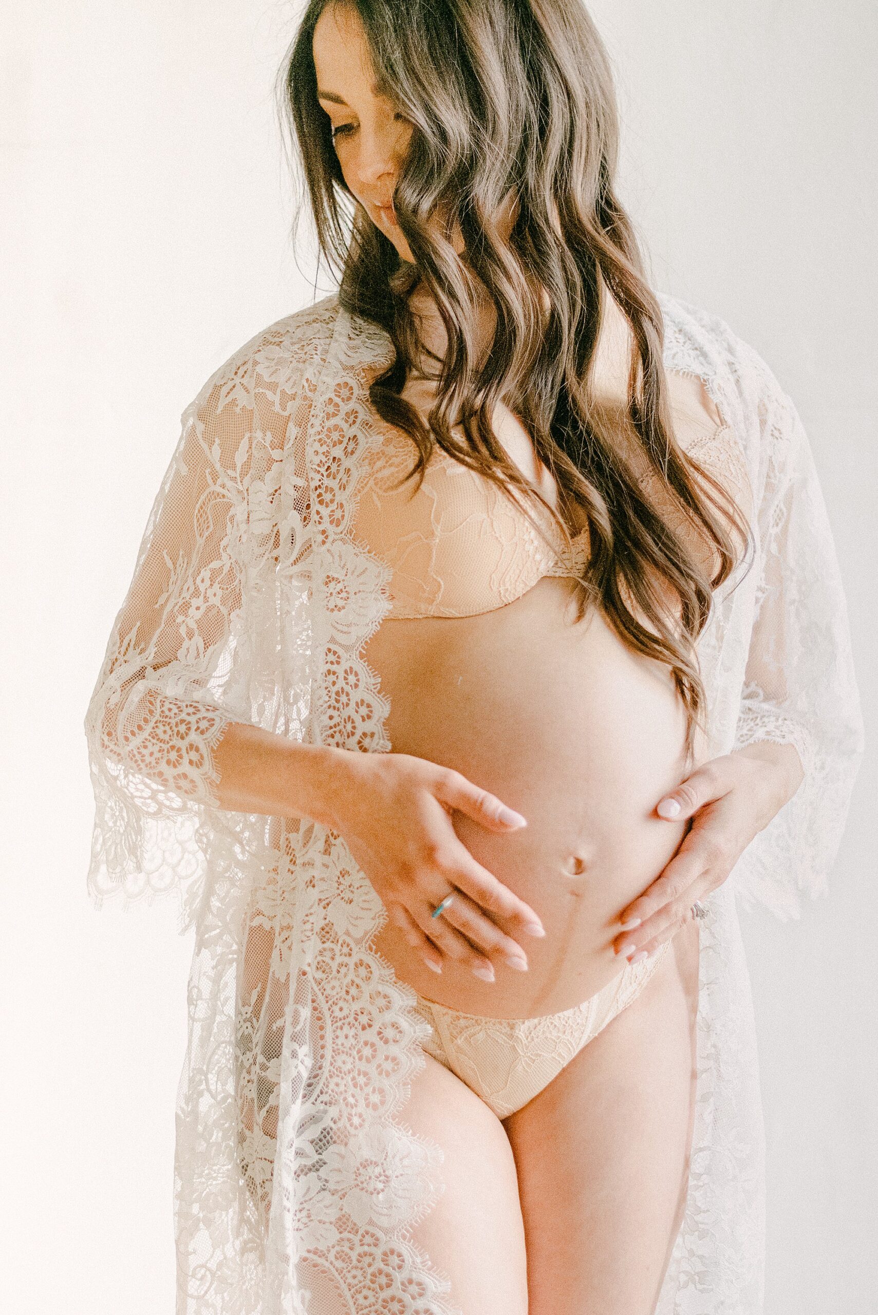 Intimate maternity boudoir photo of mom in white lace robe. She has both hands on her belly while looking off to the side with her hair on her face.
