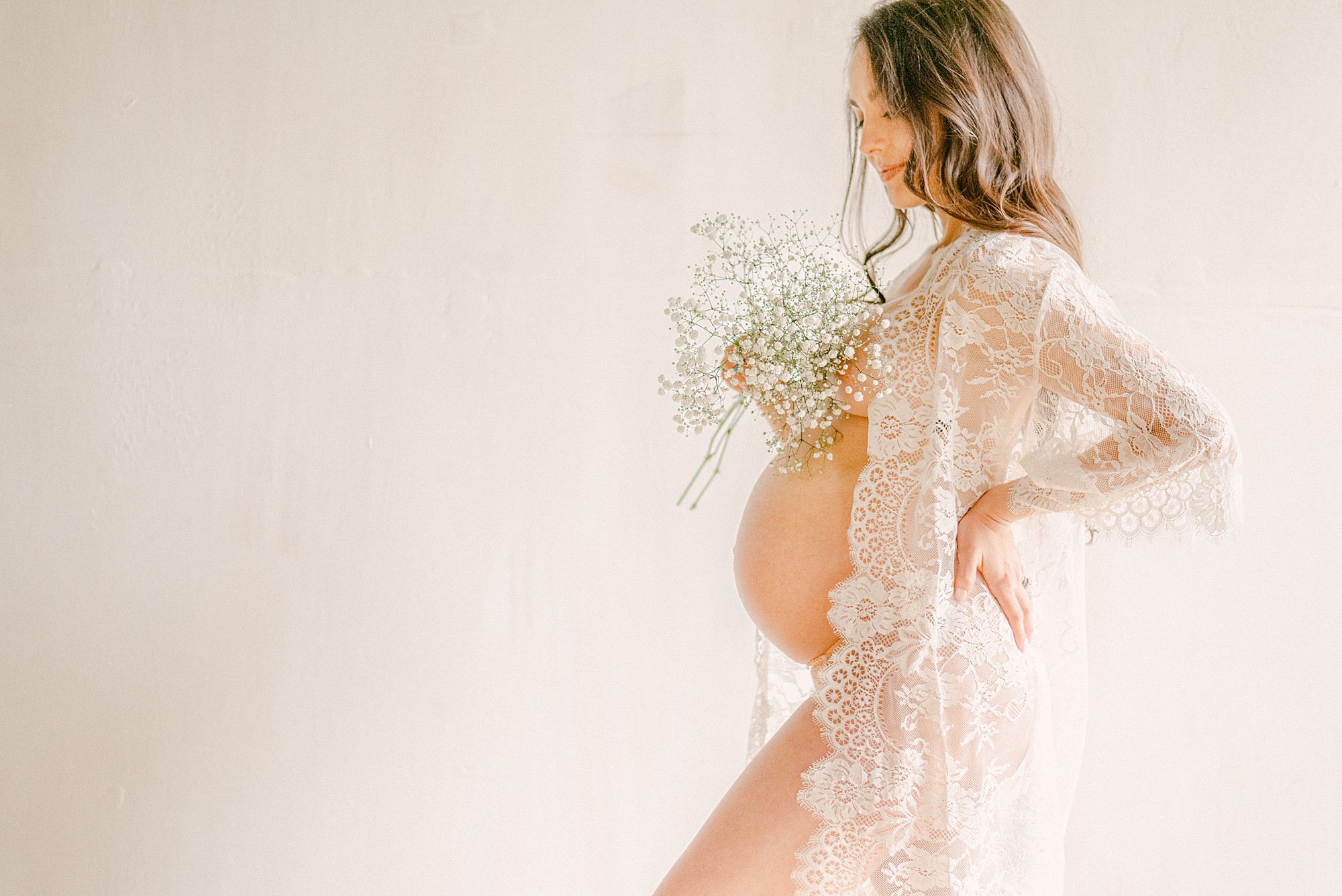 Expecting mother in lace robe and holding baby's breath. She's standing in a natural lit studio with mineral wall backdrop