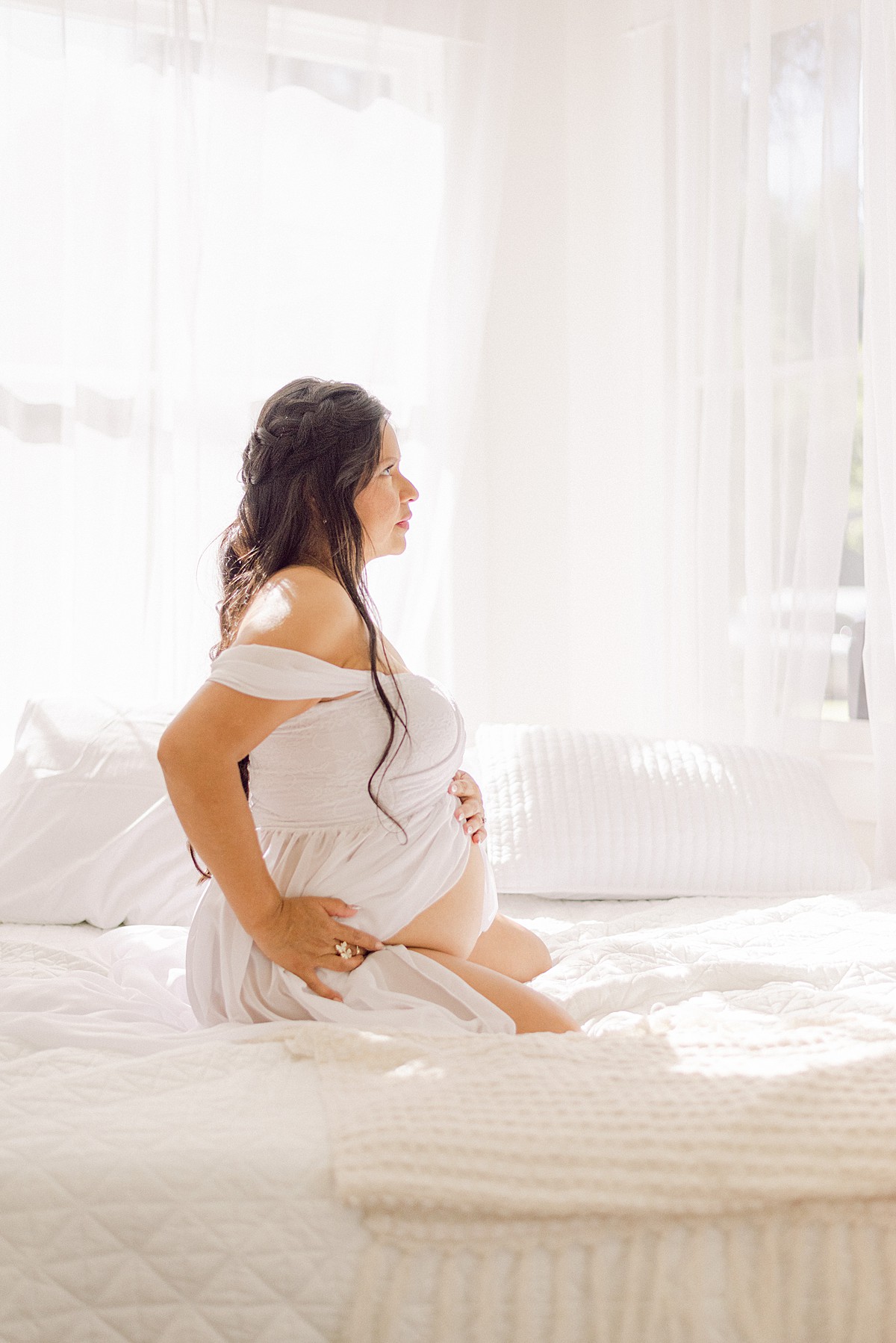Side profile of expecting mom in white dress with exposed baby bell. She is kneeling on a white bed holding her baby bump.