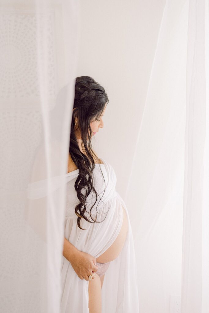 Pregnant mom standing in sheer curtains in a maternity boudoir outfit of white chiffon with baby bump exposed.