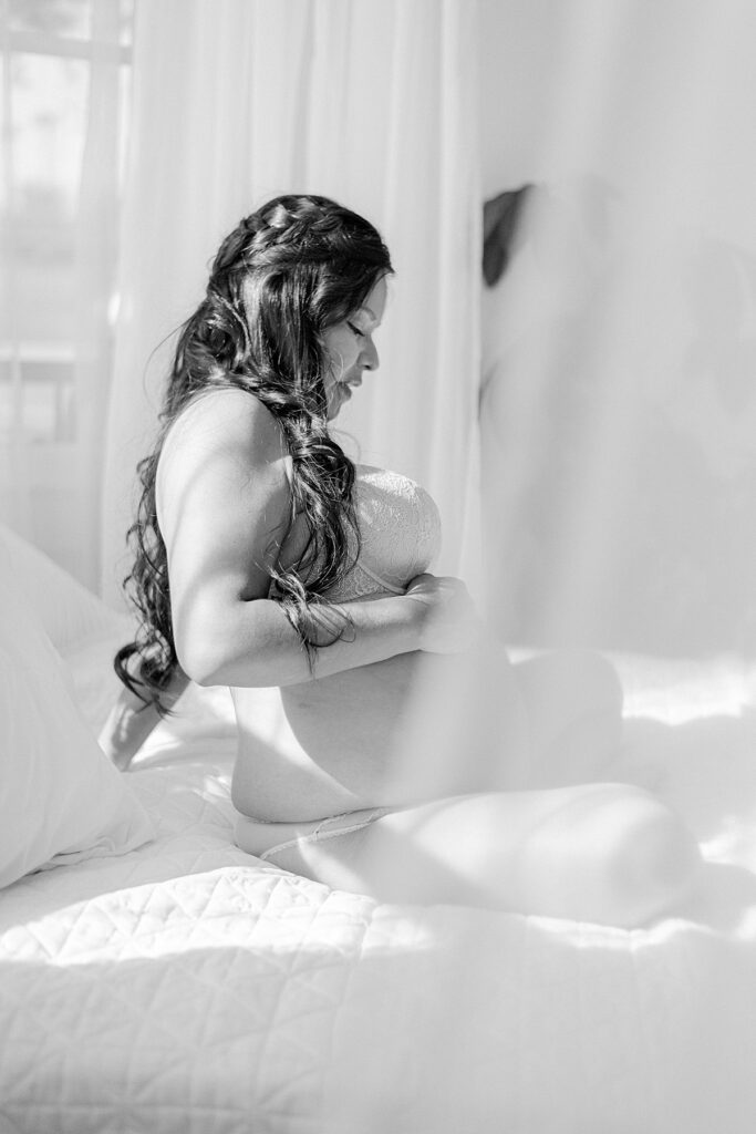 Expecting mom in blue lace lingerie while sitting on bed with hand on baby bump. She's sitting on a bed in natural lit studio surrounded by lace curtains.