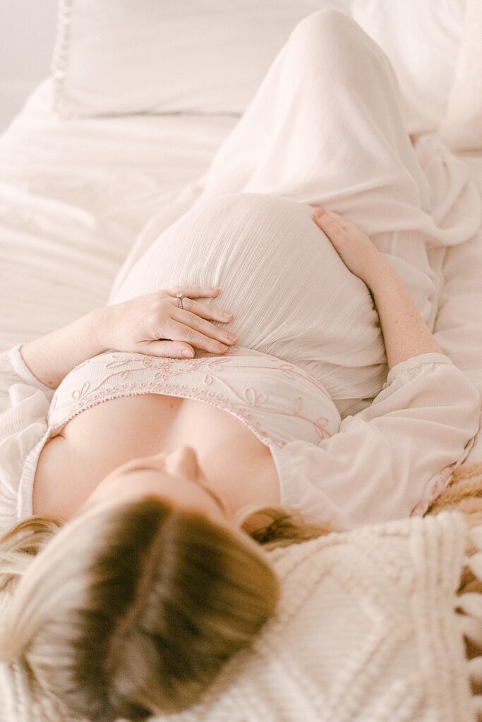 Expecting mom in white flowy dress laying on neutral decor bedding while holding her baby bump.