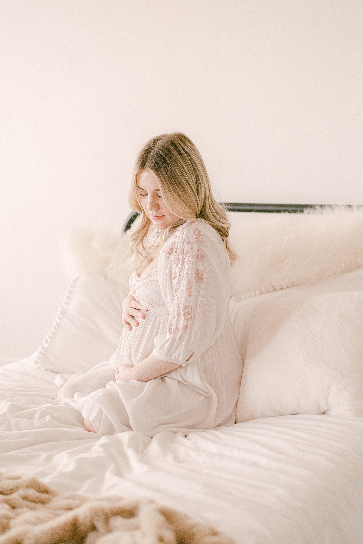 Expecting mom in white flowy dress posing for maternity photos on neutral decor bedding.