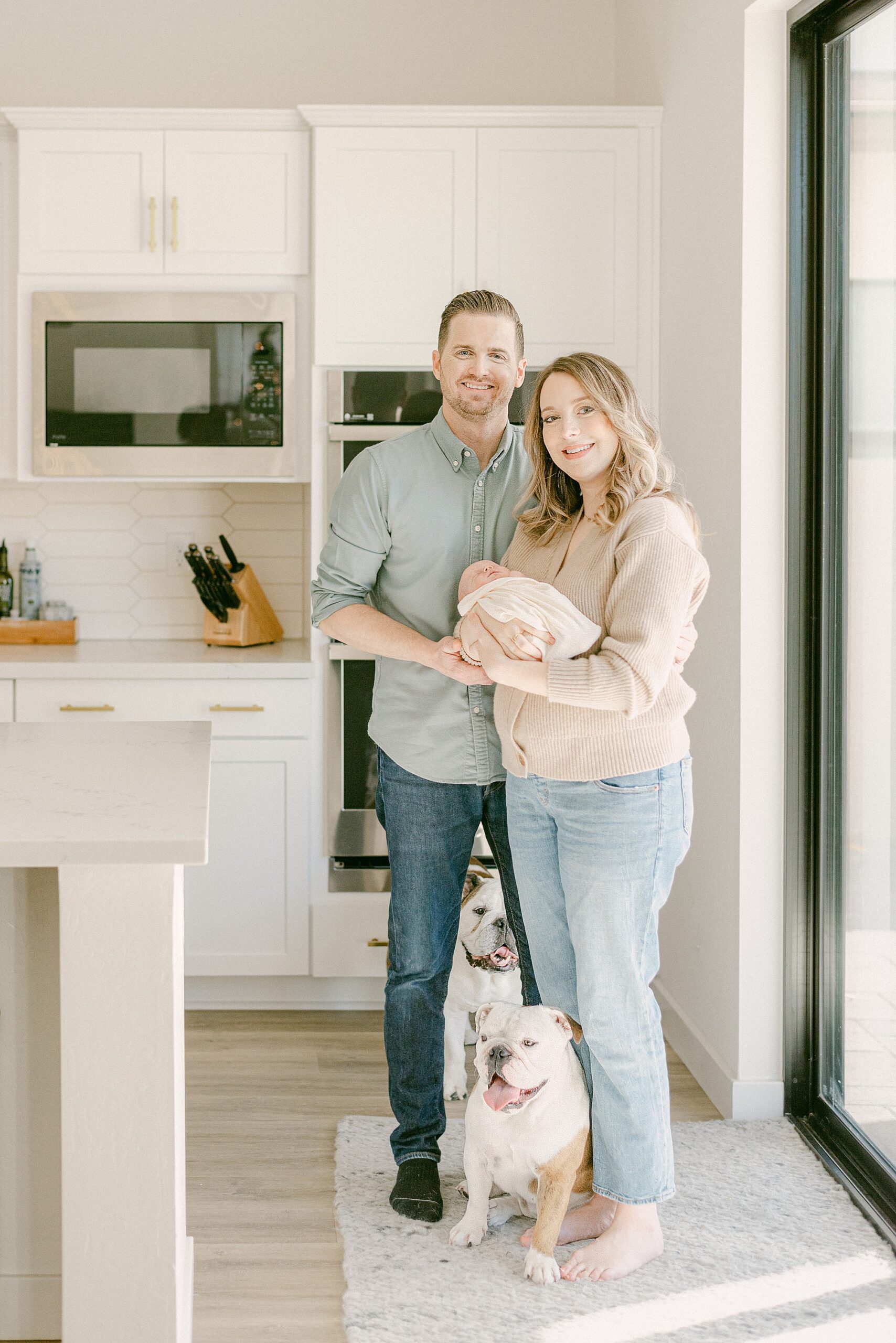 New parents standing in kitchen with baby in arms and two English Bulldogs. They were prepared on when to take newborn photos.