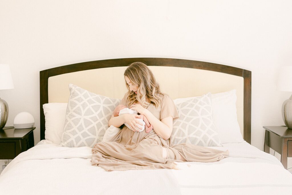 New mom wearing mauve dress while sitting in bed holding newborn baby. 