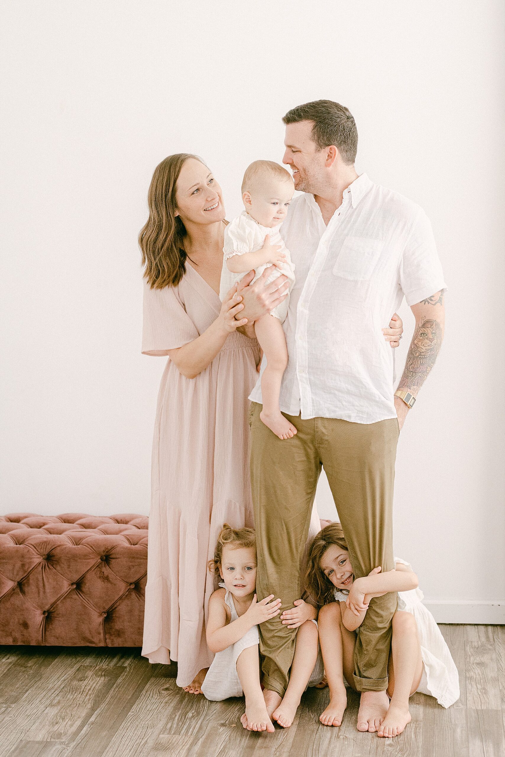 Gilbert family of five wearing neutrals and blush pink tones for family photoshoot. Mom and dad are looking at each other while hold 1-year-old daughter. Two toddler girls are at dads feet smiling at camera.