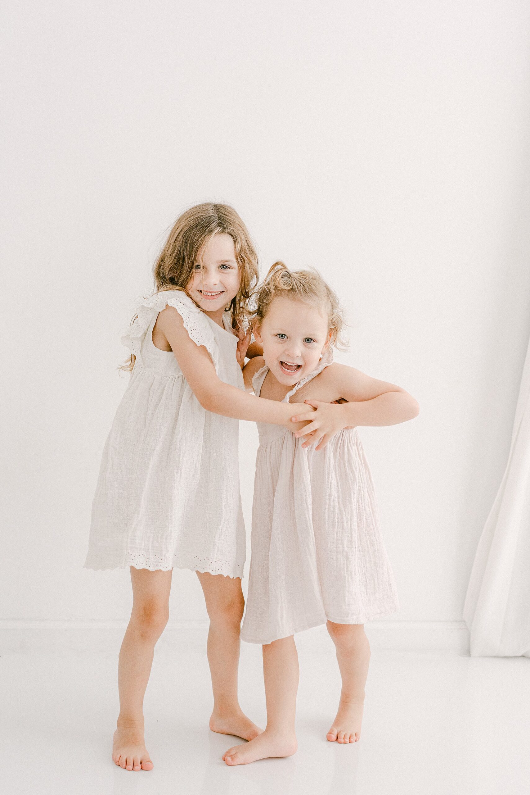 Two toddler girls dressed in white and lilac linen dresses hugging each other with big smiles