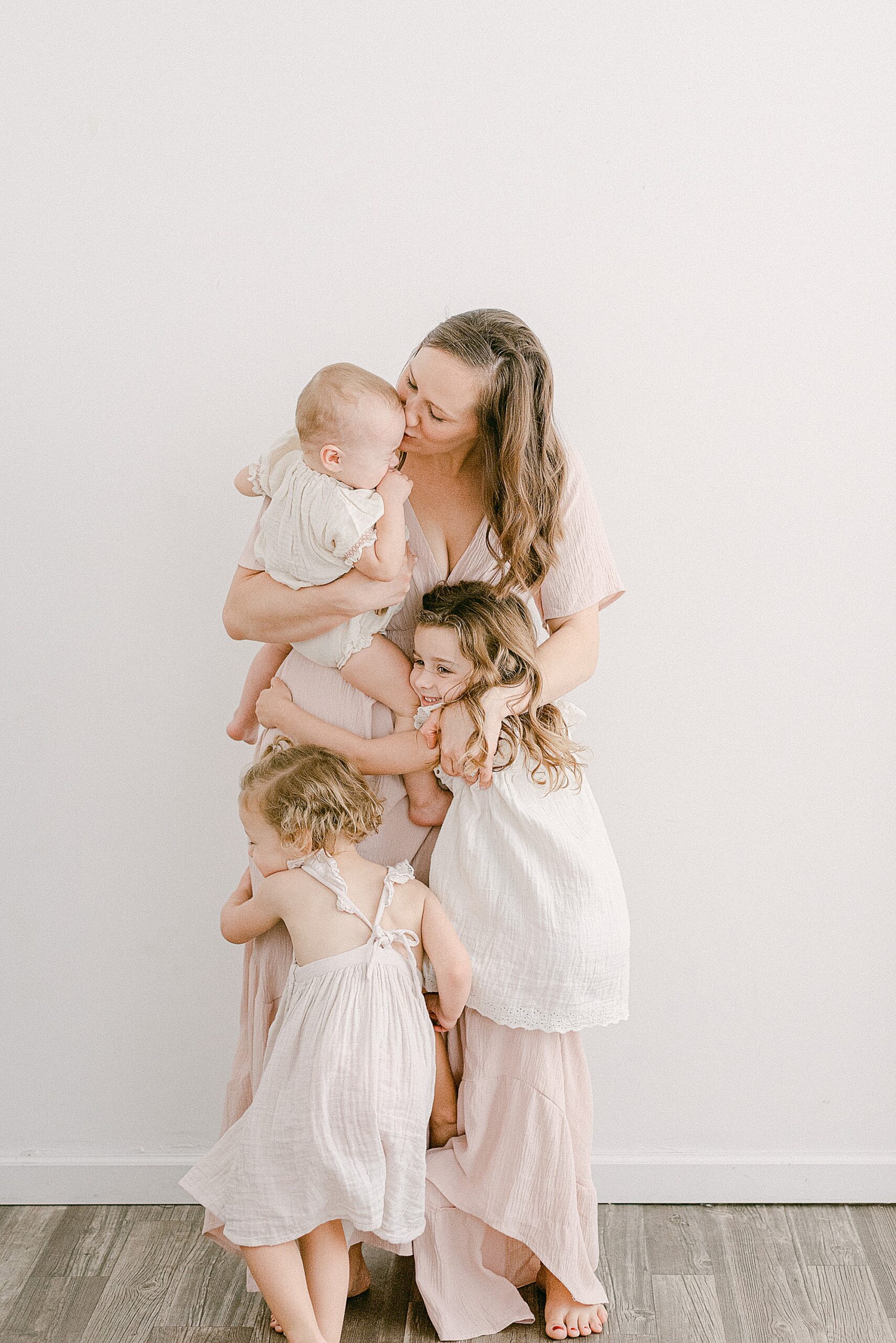 Gilbert mom being hugged by three toddler girls. They're all dressed in white and blush pink dresses for family photoshoot.
