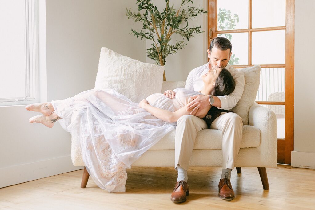 Couple knew when to take maternity photos showing mom's baby bump while sitting on cream. sofa. expecting mom is laying horizontal cradling belly and dad is kissing her forehead.