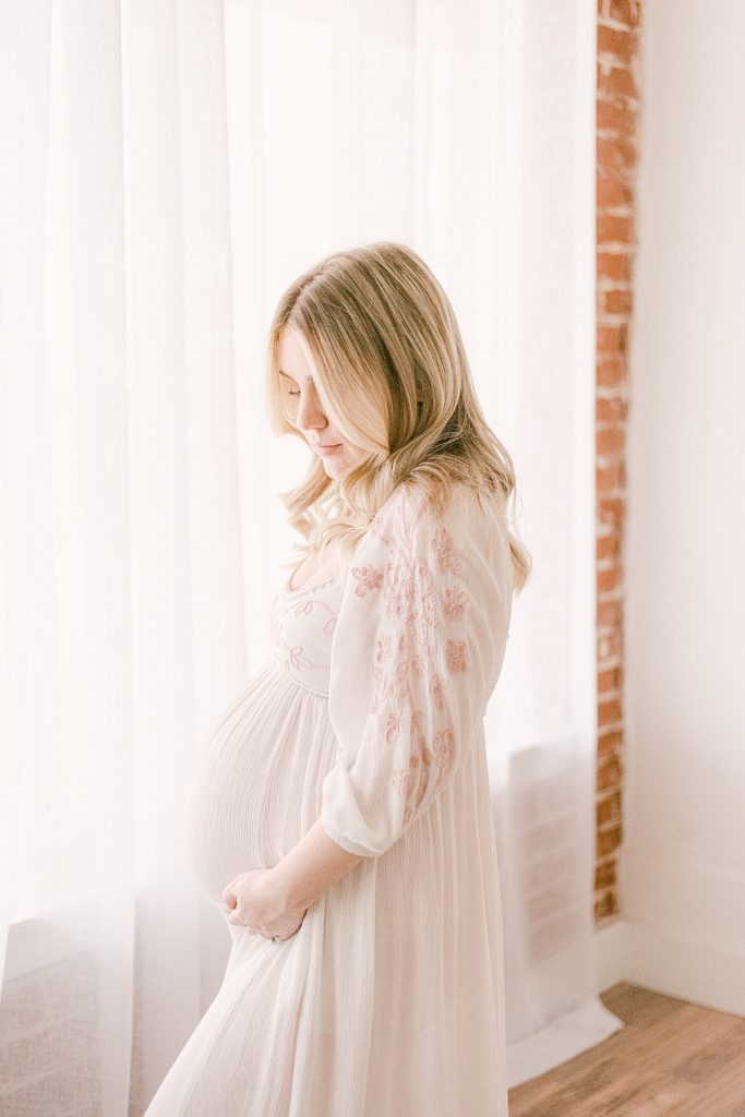 Mom in 2nd trimester dressed in neutral cottagecore dress with pink details on sleeve. Mom is snuggling baby bump while standing next to a window in studio with brick wall
