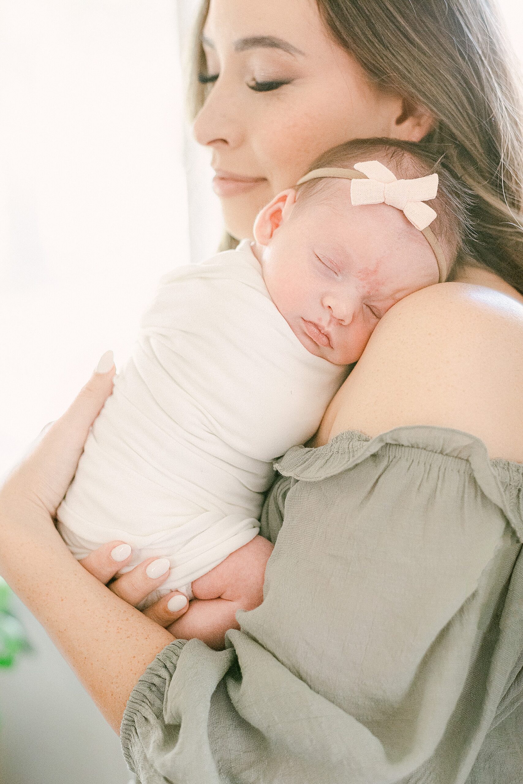 Mom with closed eyes and soft smile is holding newborn baby girl with her head resting on her shoulders.