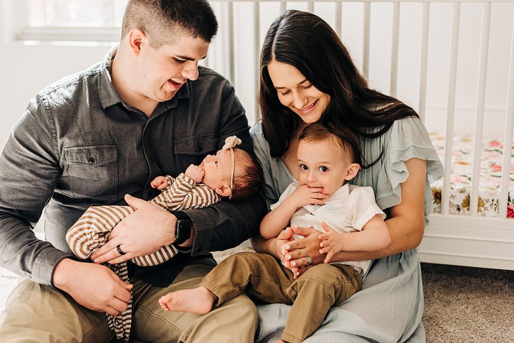 family of four sitting on floor in front of white crib for in home newborn photos