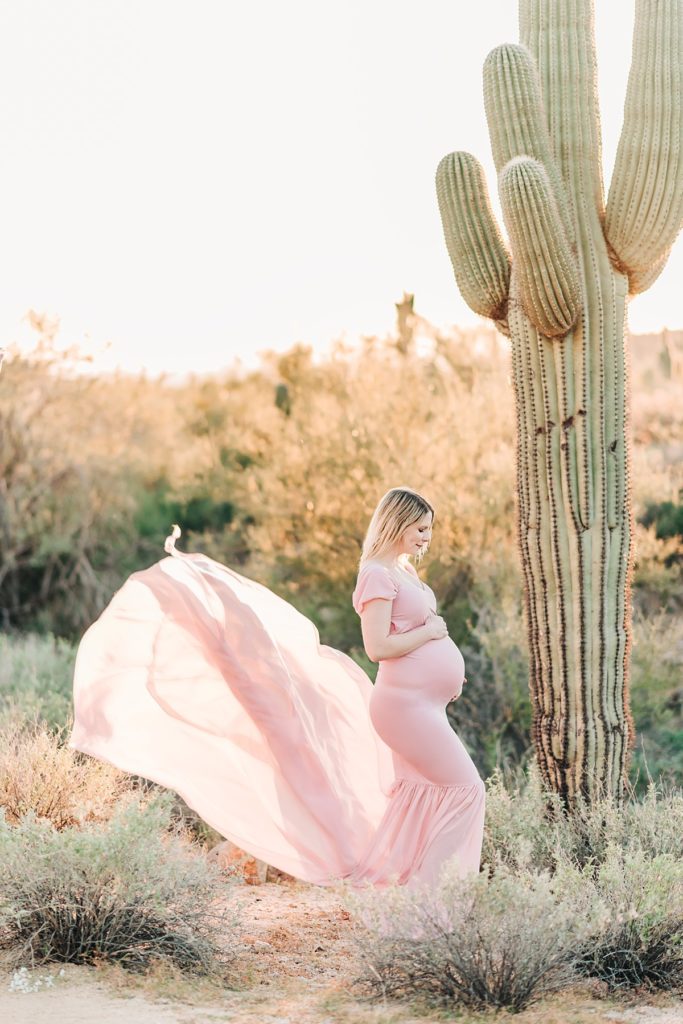Woman holding baby bump in blush pink flowing dress while standing next to saguaro cactus