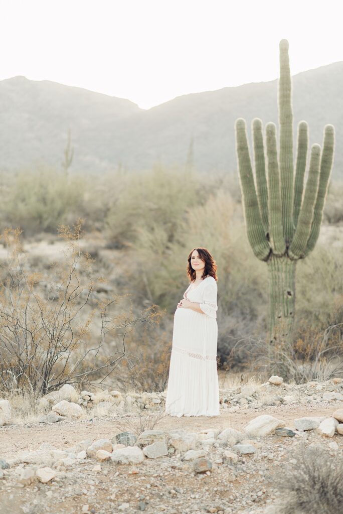 mom in white dress has hands on baby bump while standing in golden hour light by saguaro and mountains in background