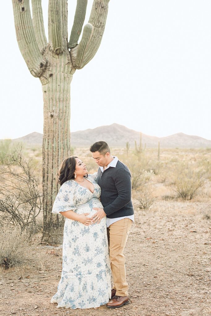 expecting couple in blue and neutral themed clothing standing next to saguaro in desert. theyre cradling the baby bump