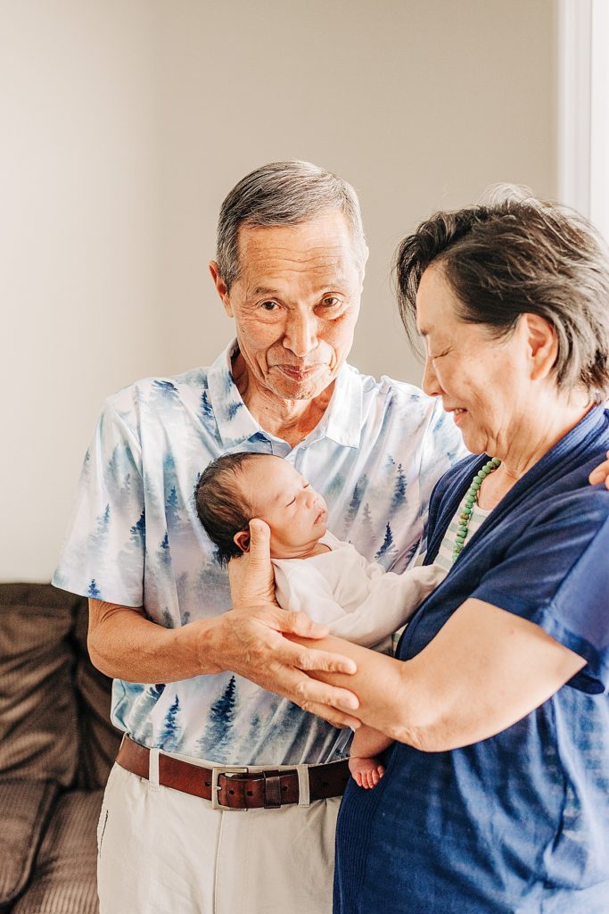 Grandparents holding newborn grandson joyfully smiling together. Grandfather is smiling at camera. Grandmother is smiling at grandson.