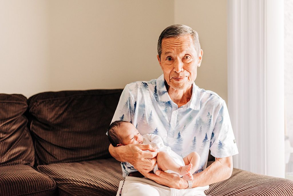 Grandfather holding newborn grandson while sitting on couch. Grandfather is looking directly at camera with proud and endearing look on his face.
