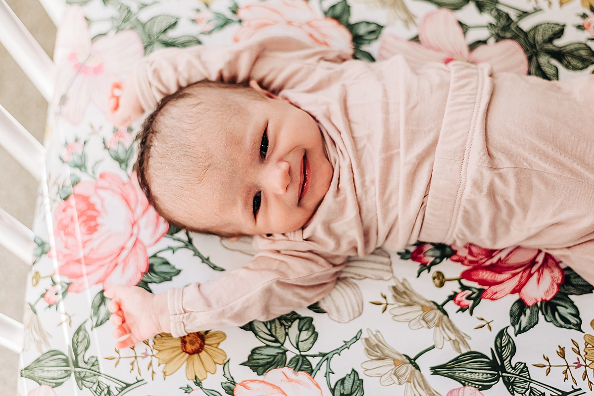 Newborn baby girl in blush pink pajama set. Baby is in a white crib with a floral patterned crib sheet.
