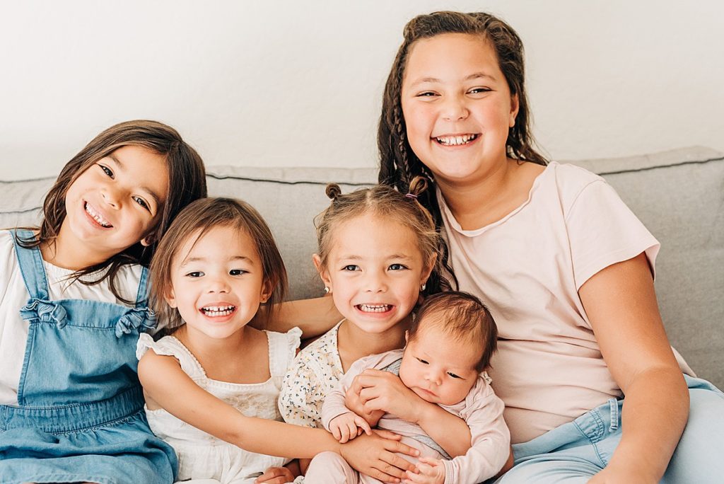 Five daughters aged from newborn to early teen all together on couch for lifestyle newborn photos in home. 