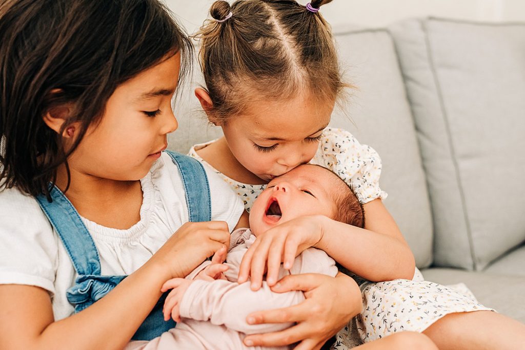 Toddler girl kissing newborn baby sister on forehead. Newborn baby is yawning.