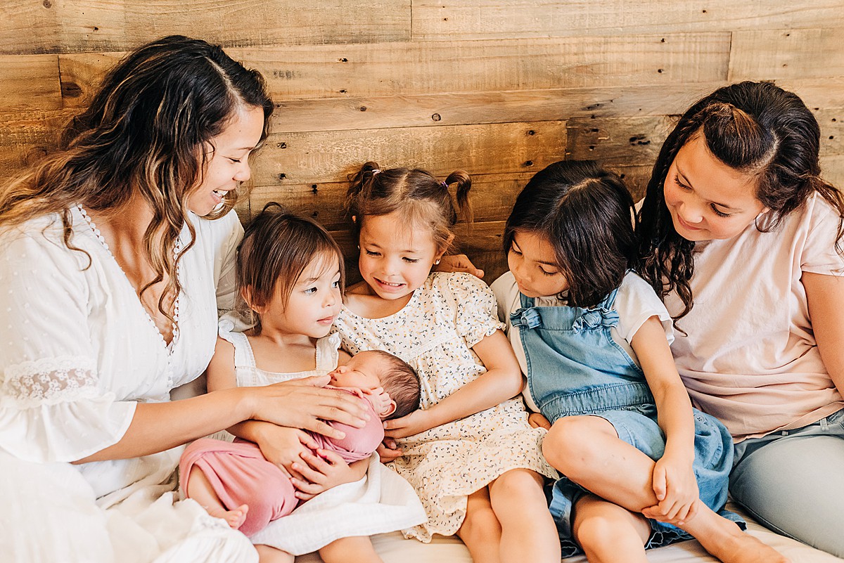 Mom and 5 daughters cuddled on bed for lifestyle newborn photos in home.