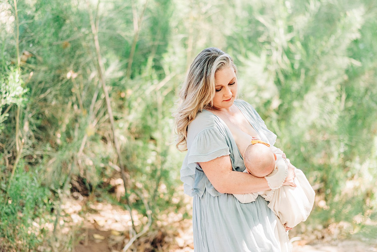 Mom and baby are surrounded by green bushes while mom is standing and holding 6 month old breastfeeding baby. Mom is in a loose baby blue dress and baby in neutral colored onesie.