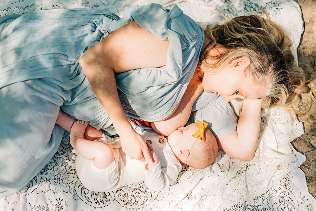 Mother side laying on blanket while breastfeeding baby. Mom is wearing baby blue flowy dress and baby in a neutral colored onesie.
