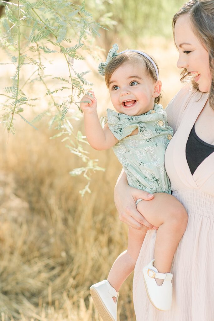 Toddler girl in mom's arms grabbing stem from a tree
