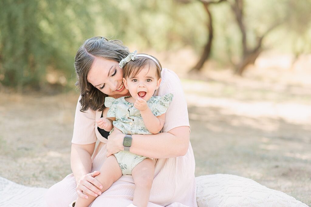Mom holding toddler in lap. Toddler has a piece of grass she's holding in her mouth.
