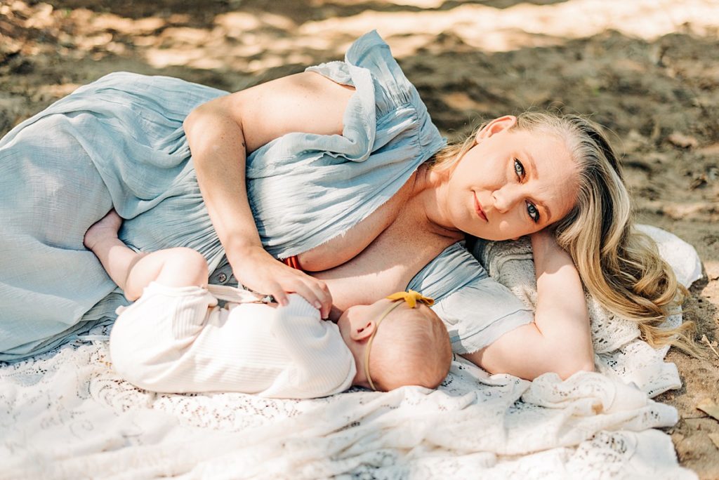 Mom in baby blue flowing dress is side laying on a neutral colored blanket. Mom is breastfeeding baby girl in neutral colored onesie. 