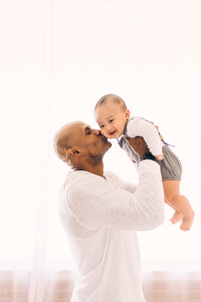 Dad lifting 6 month baby boy in the air and kissing on cheek. Baby boy is dressed in gray/blue overalls with long-sleeved white cotton onesie