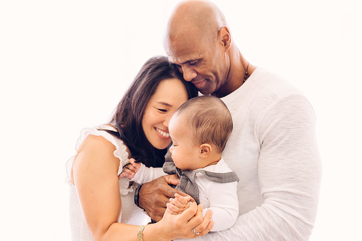 Light and airy baby photo of a couple holding a 6-month baby boy. Baby is wearing blue/grey overalls with white long-sleeved onesie.