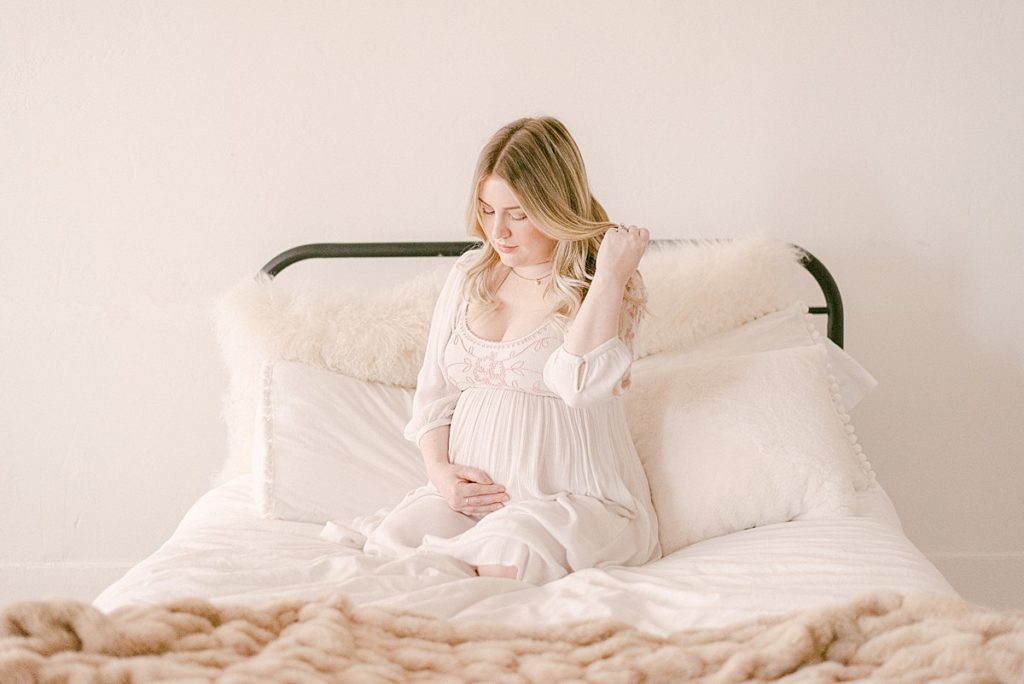 Mom-to-be in flowy white dress on neutral decorated bed. She's running her hand through her hair and snuggling her baby bump.