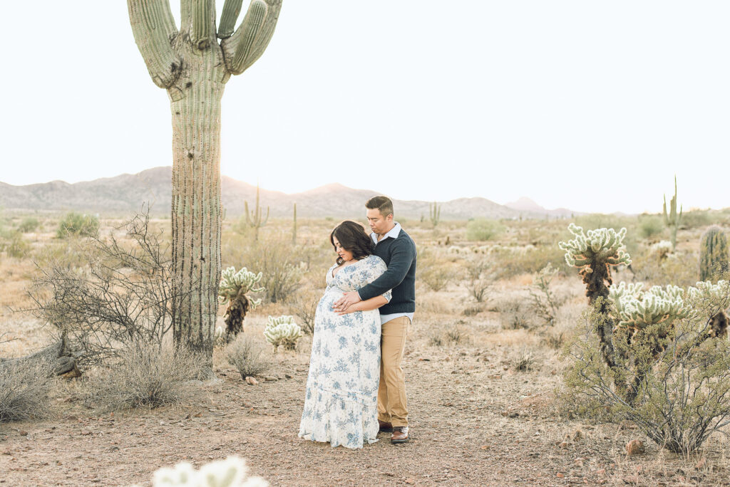 Couple expecting a baby are embraced while standing in desert cactus scenery in Scottsdale, AZ