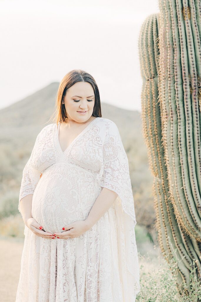 Mother to be in white lace gown holding baby bump. She's standing next to saguaro cactus in scottsdale, arizona.