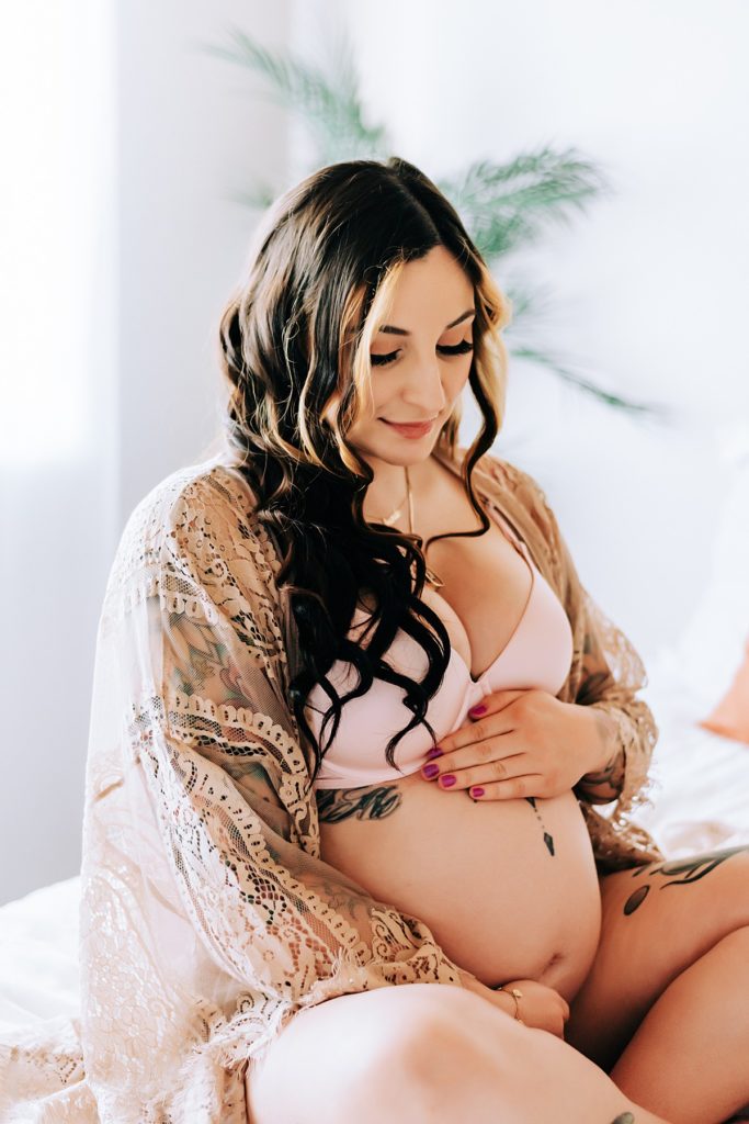 In-studio Maternity boudoir session of mom in lace robe. She is holding baby bump while sitting on bed in neutral boho decor.