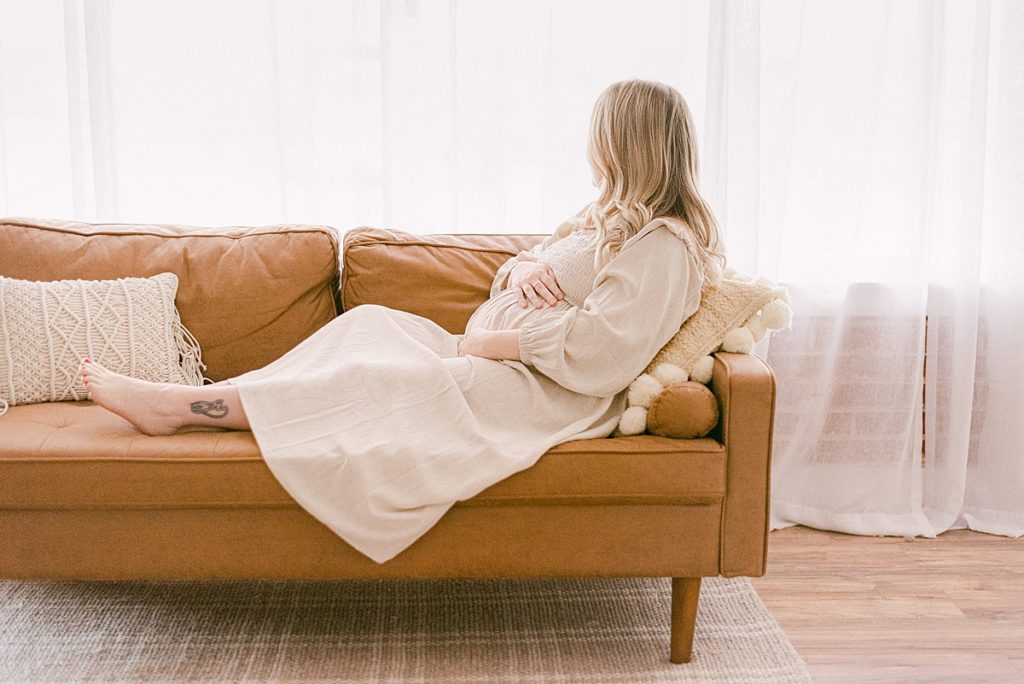 candid intimate maternity session in light and airy studio. mom is holding baby bump and wearing cream flowy dress by rachel pally. mom is sitting on tan leather couch