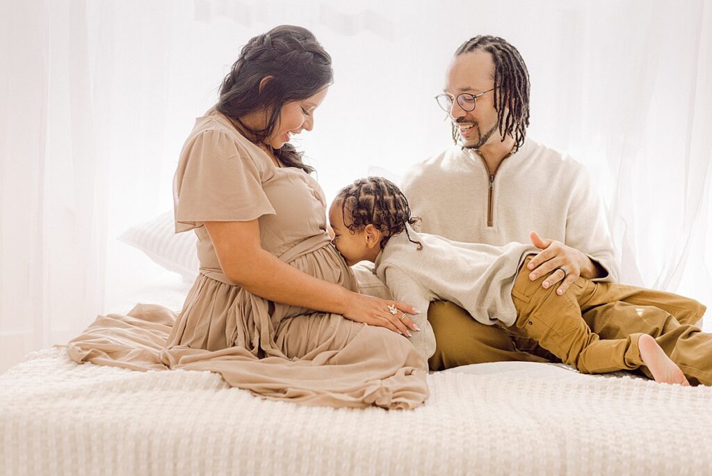 Mom and Dad on bed with toddler son kissing mom's belly for maternity photos