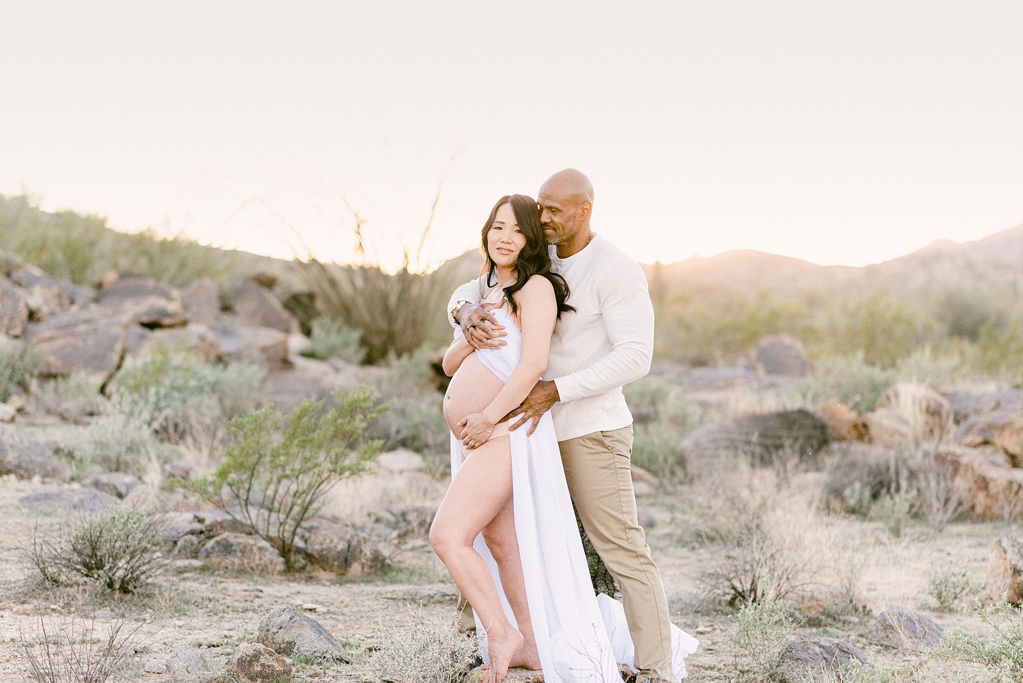 couple embraced in arizona desert for maternity boudoir. Pregnant woman is weari chiffon maternity fabric
