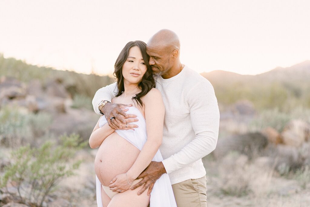 man snuggled behind woman who is draped in chiffon for a desert maternity photoshoot