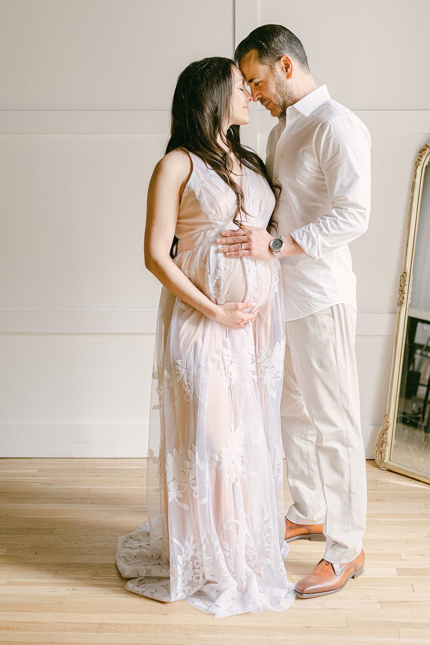 Expecting couple wearing elegant neutral clothing with foreheads together. Their hands are on baby bump