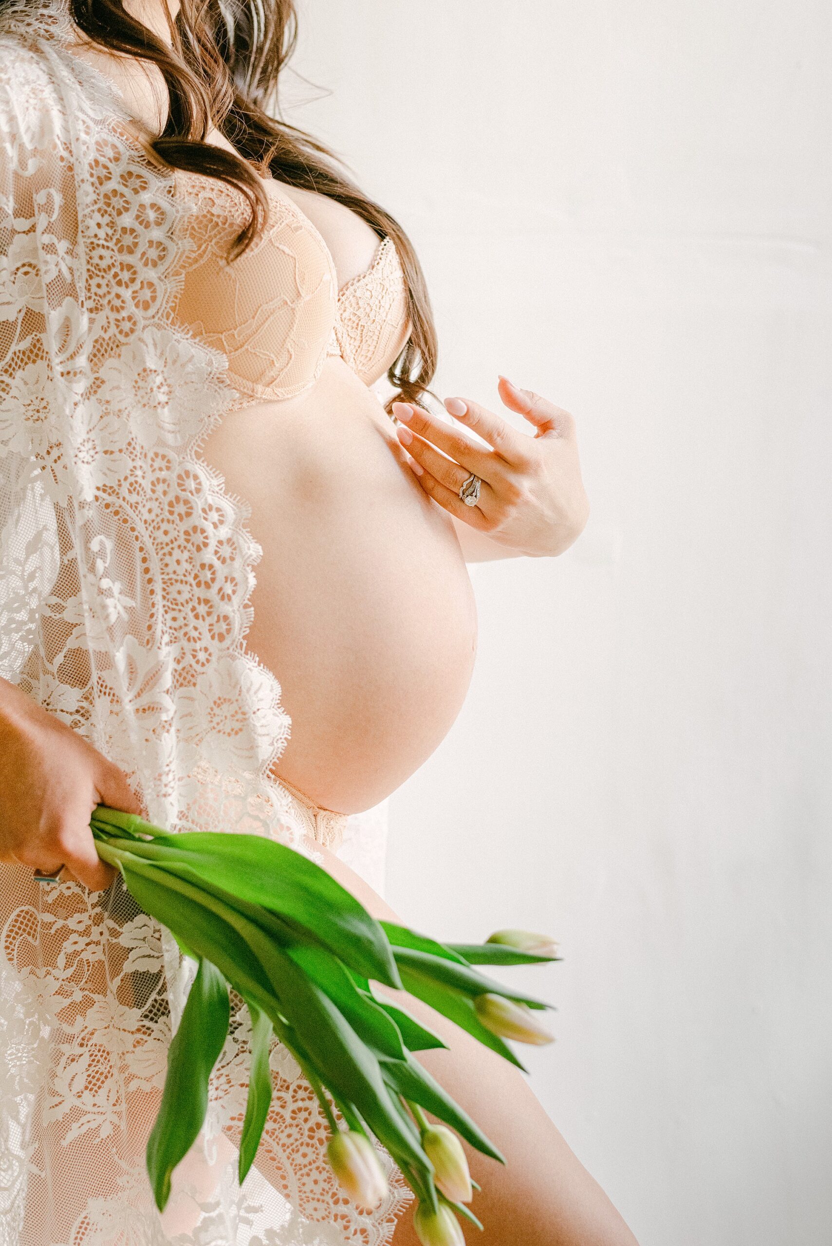 Expecting mother holding tulips next to baby bump. She's wearing a white queen anne lace rob with fingers tracing baby bump