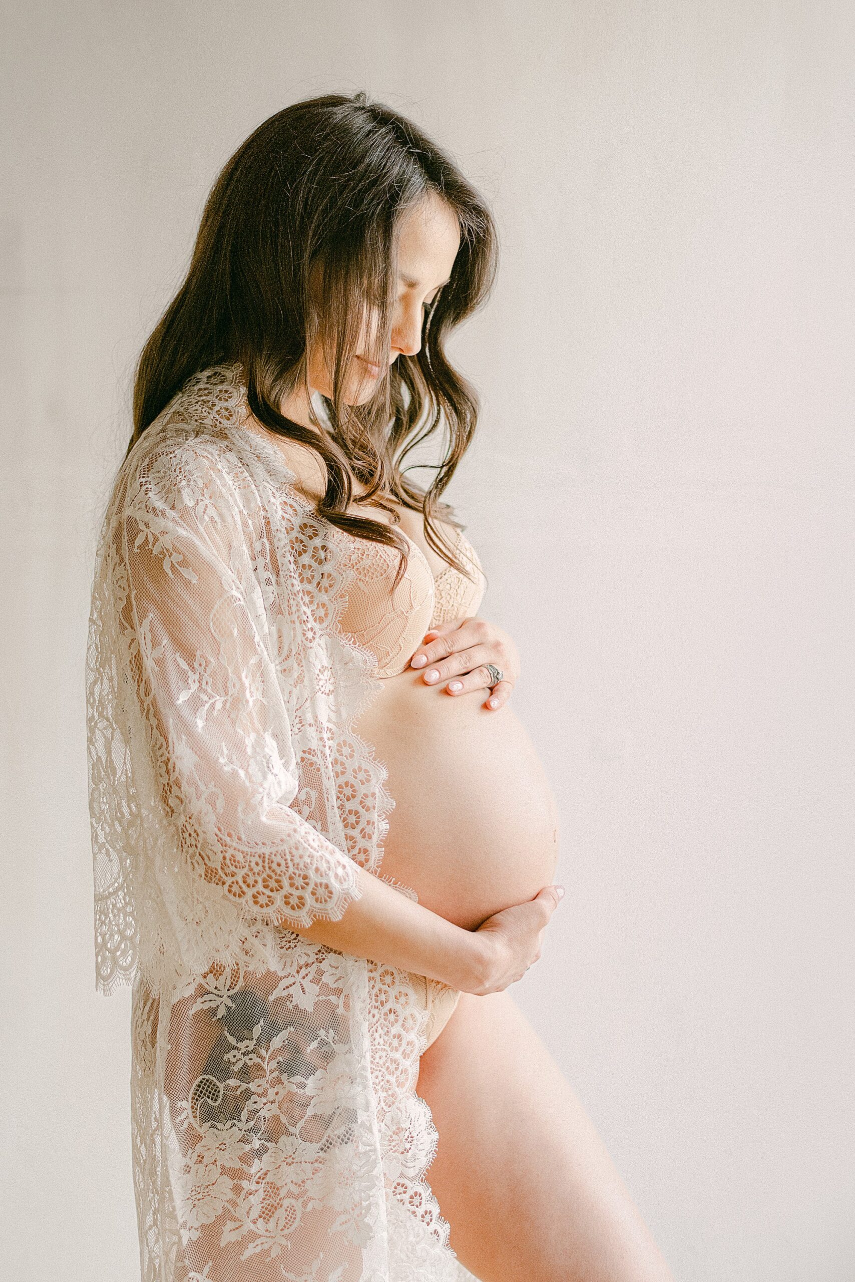 Side profile of expecting mom in white lace robe holding baby bump
