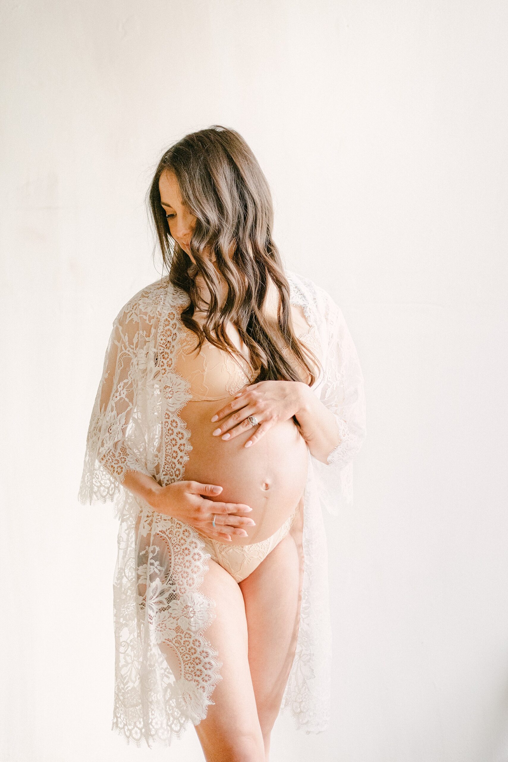 Pregnant mom holding baby bump while wearing pink lingerie and white lace robe. She is facing her shoulder and hair falling over face.