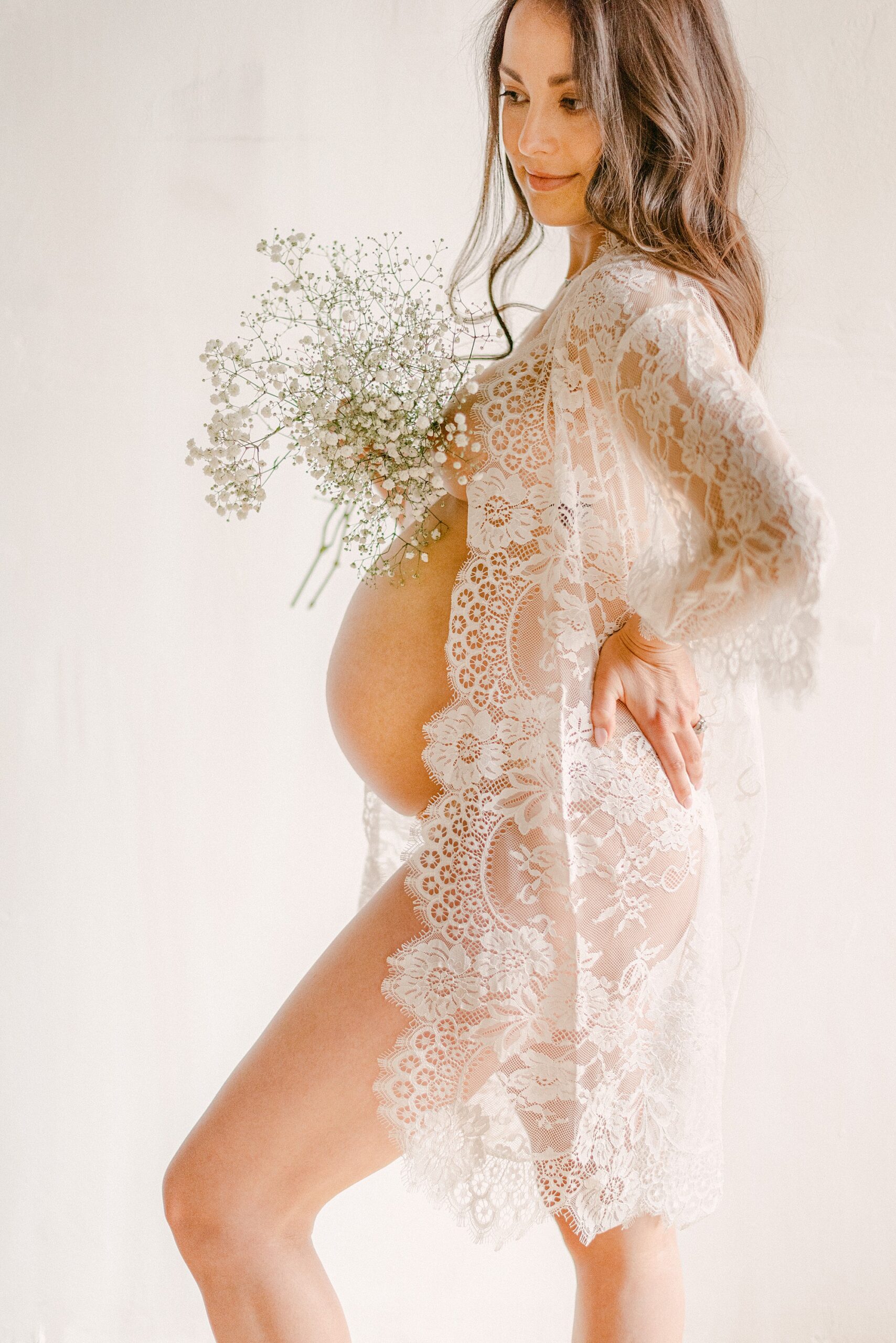 Mom to be in white lace robe holding baby's breath flowers above baby bump.