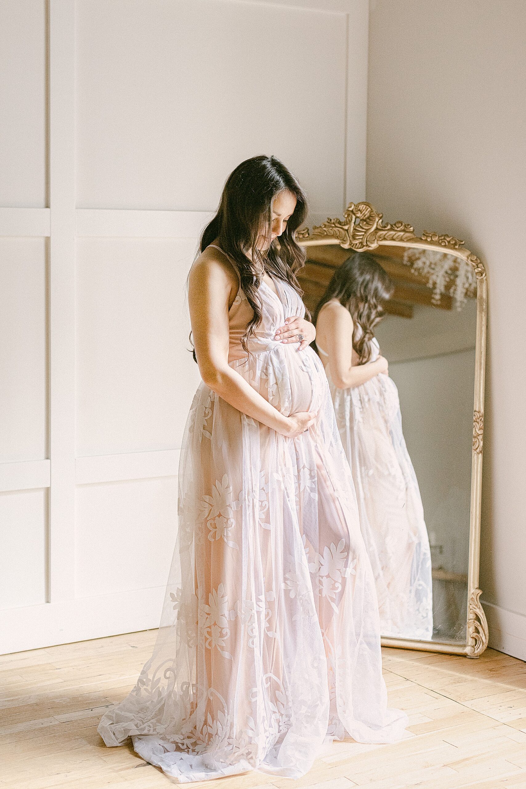 Expecting mom in elegant white lace dress in front of anthropologie gold mirror holding baby bump. She's standing in-studio with board and batten walls.