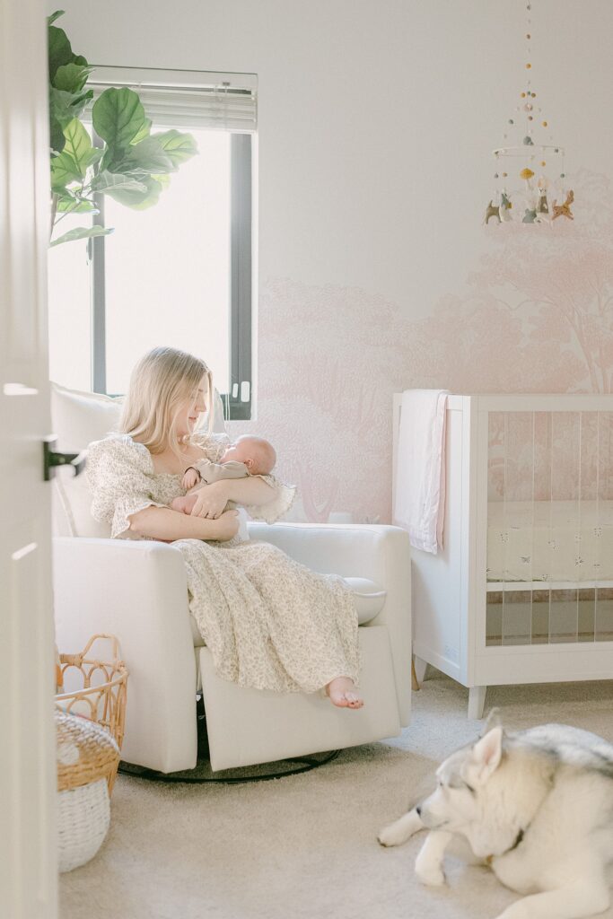 New mom in neutral and pink decorated nursery. Mom is sitting on glider while holding baby. Siberian husky is at her feet.