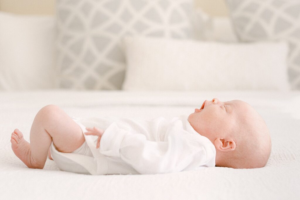 Newborn baby in white onesie yawning while laying on neutral colored bed