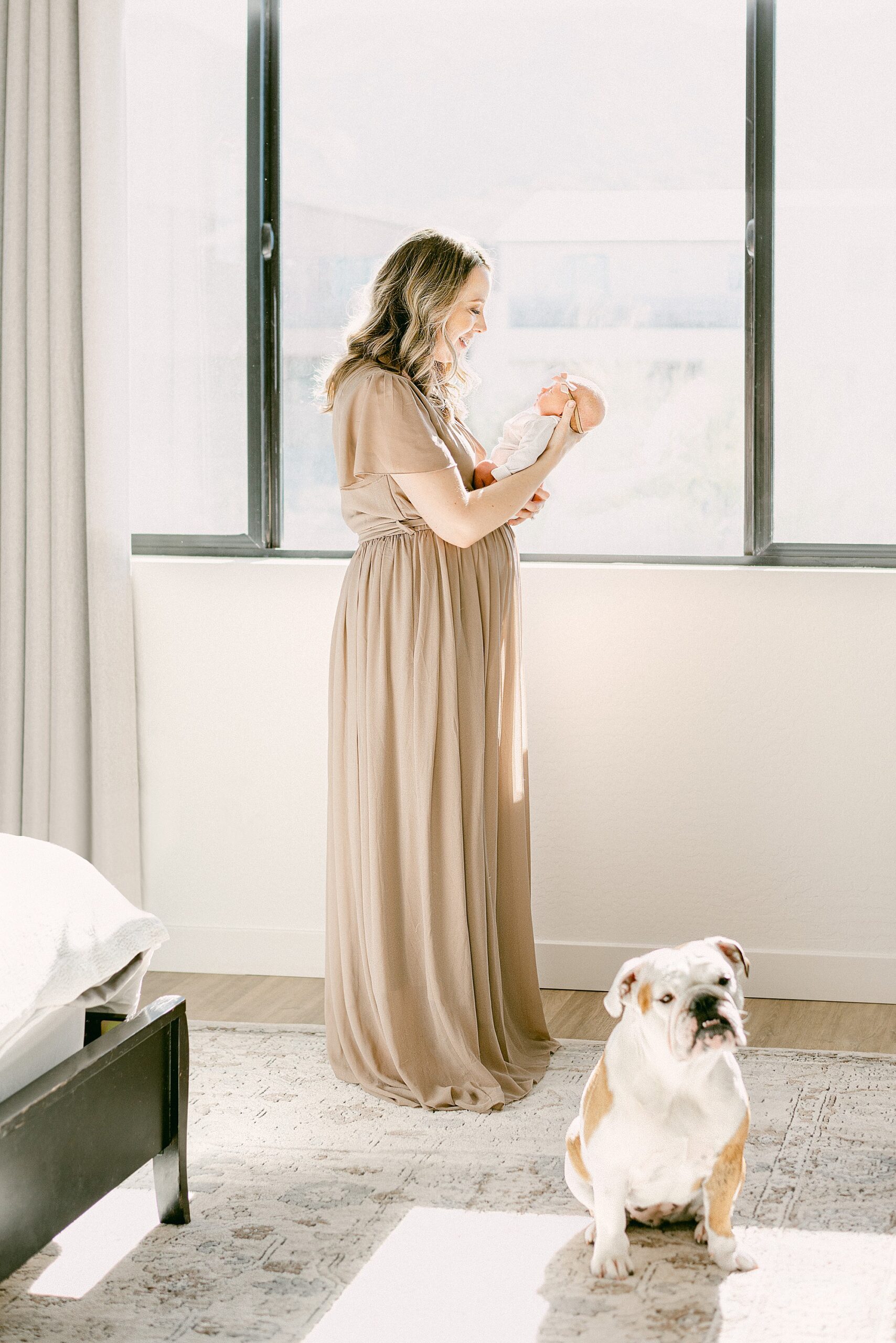 New mom wearing mauve gown standing next to window while holding newborn baby face to face. English Bulldog is sitting next to her feet while looking at camera.