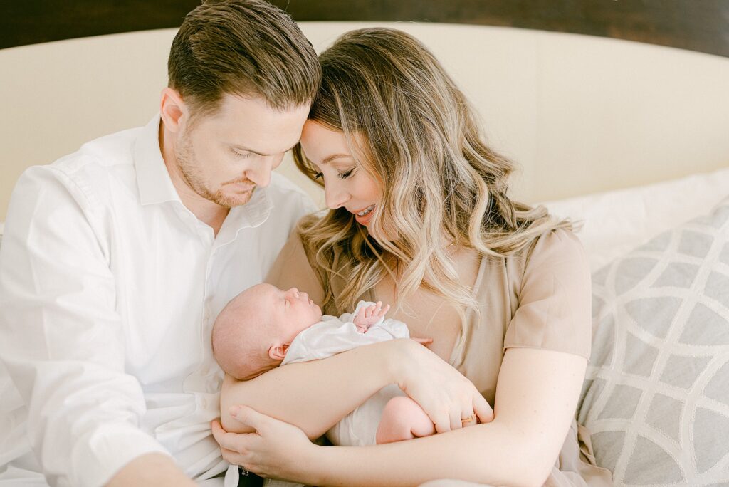New parents cuddled on bed holding newborn baby for in-home photos